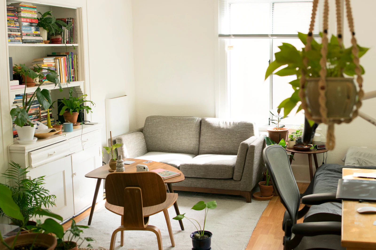 Compact apartment living area with a small sofa, round table, plants, bookshelves, and a workspace edge, shown as small-space inspiration.
